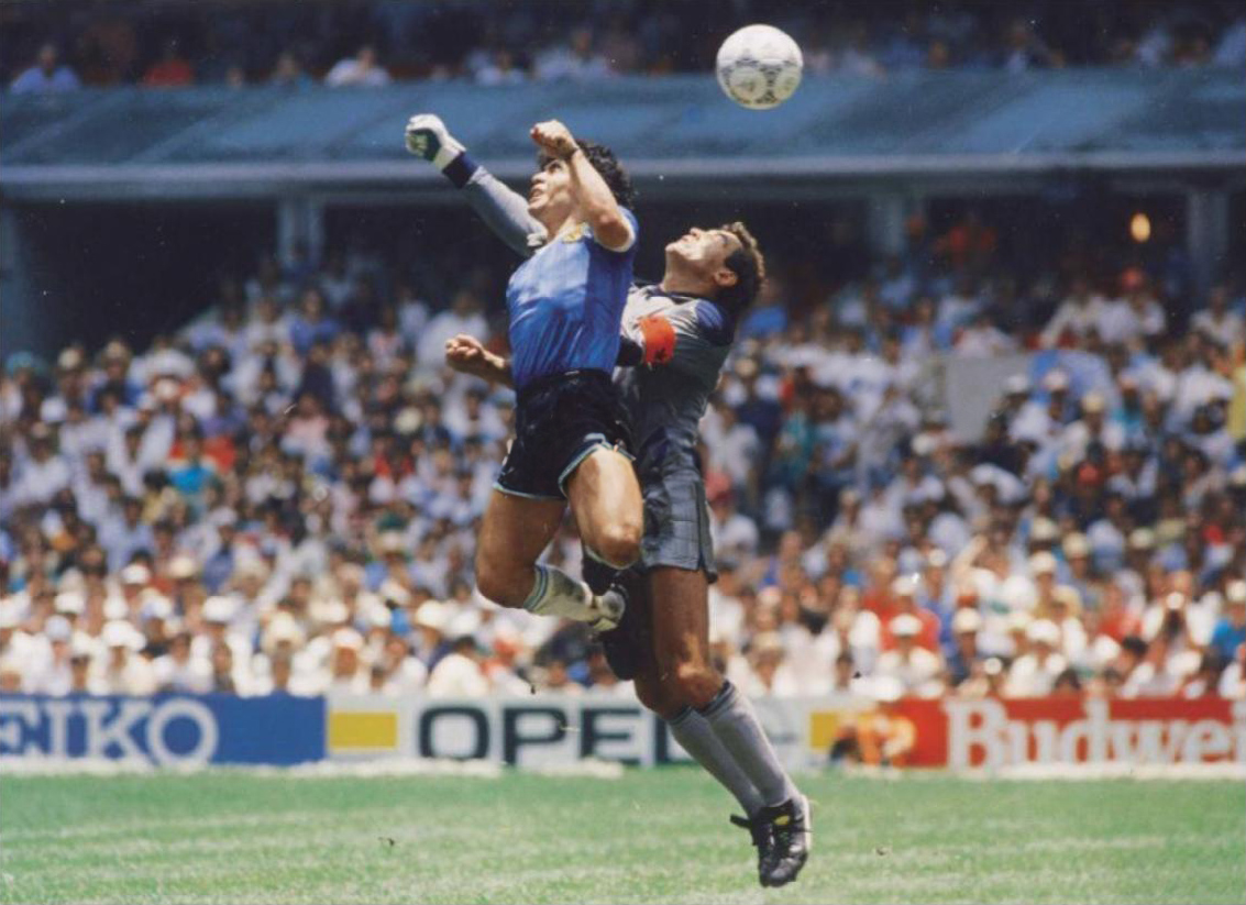 Maradona salta ao lado do goleiro Peter Shilton e toca a bola com a mão esquerda no lance da Mão de Deus, no Estádio Azteca em 1986